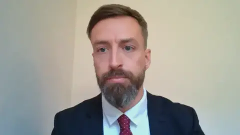 Liam is looking at the camera through Zoom with a plain white background. He has well-kept short brown hair in a quiff, and a full beard that is greying near his chin. He is wearing a black suit, white shirt and red spotted tie.