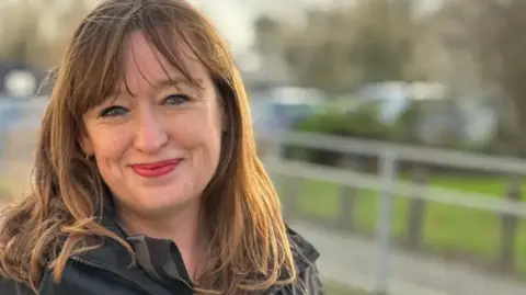 A woman with brown hair is stood outdoors looking into the camera.