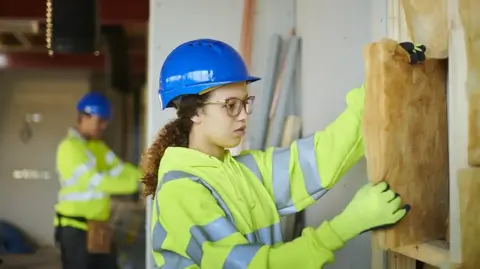 Getty Images A woman in a hard hat and fluorescent jacket handles insulation inside a building. A man is in the background in a similar jacket and hat.