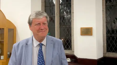 A mean wearing a white top and blue tie. He has a light blue blazer on. Behind him are white walls with large glass windows.