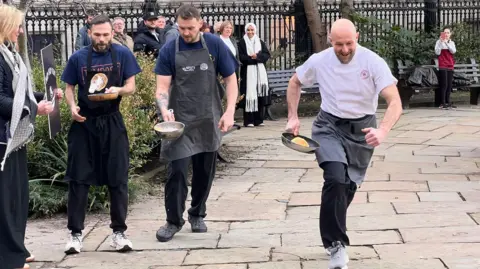 Three chefs wearing aprons flipping pancakes in frying pans as they run around the gardens of Liverpool Parish Church