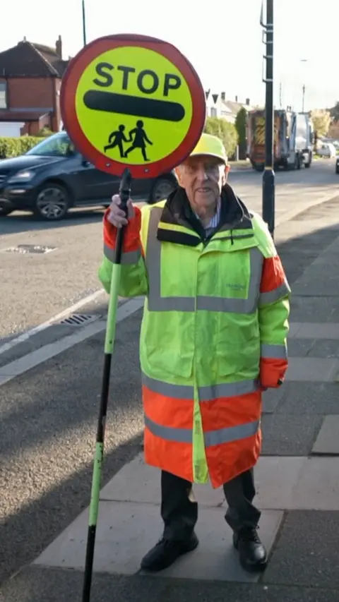 An elderly lollipop man, stood smiling in his hi-vis coat and stop sign.