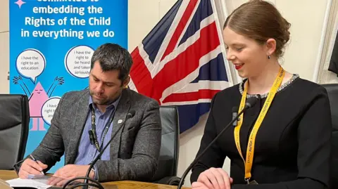 Councillor Leigh Frost with Rosie from the Cornwall Youth Council sat beside him. He is signing a document and is wearing a grey blazer. 