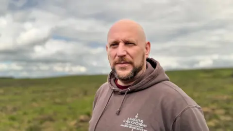 BBC A bald headed man with ginger, dark brown and grey facial hair looks at the camera as he stands in the mostly green peatland. He is wearing a brown hoodie which has white writing in the corner.