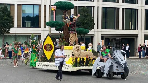 BBC A float featuring a lion, ostrich, colourful bird, African-style shields and a tall tree. It is being towed by what looks like a ride-on mower that is dressed up to look like a rhino. A woman walks behind the float wearing a bright green skirt, top and a headdress which have exotic fruit on them. There are also men dressed in green carrying African-themed shields. There are spectators on the streets.