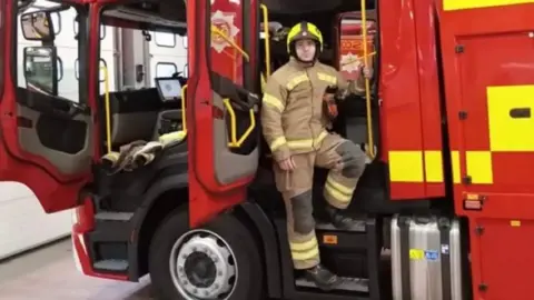 SFRS Barry Martin is standing on the steps of a red fire engine, looking at the camera. He is wearing brown fire-resistant overalls with hi-vis stripes on them and a yellow and black helmet 