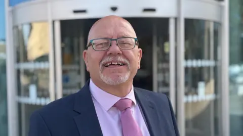 Local Democracy Reporting Service A bald headed man with glasses and a goatee smiles into the camera. He has on a dark suit with a pale pink shirt and darker pink tie. He stands in front of a large glass doorway.