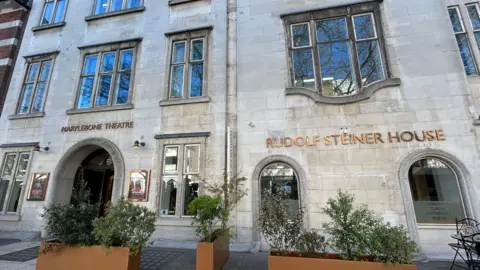 BBC/Harry Low Exterior of Rudolf Steiner House in gold writing along with a sign for the Marylebone Theatre. Plants are in front of it 