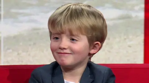 A young boy with blue eyes and blonde hair sitting on a red sofa.