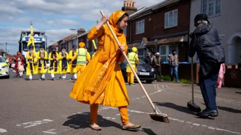 PA Media A woman in traditional orange dress, and a man in navy blue, sweep the road ahead of the Nagar Kirtan. Behind them are five men in the bright yellow outfits of the Panj Pyare (Five Beloved Ones) and a float.
