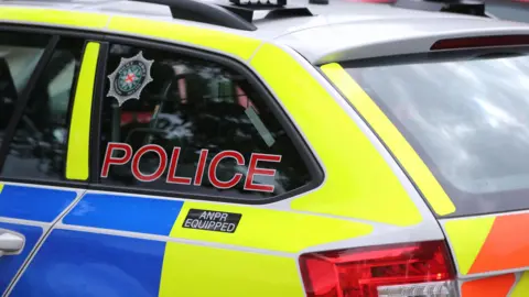 Side view of a PSNI vehicle, coloured in blue, light-green and orange. The window reads police in cpital red letters.