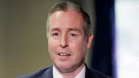 PA Media Paul Givan - a man with short hair is pictured from the shoulders up is wearing a dark suit jacket and a white collared shirt. He has his mouth open as he looks beyond the camera.