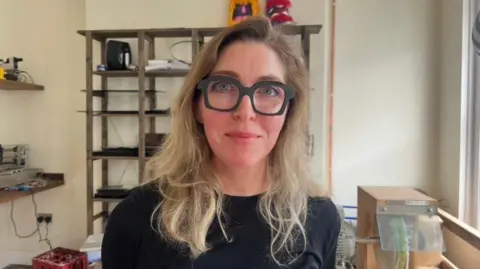 Claire Mulvenna stands in her opticians shop. She is wearing thick black rimmed glasses, and a a black jumper. She has long blond hair.