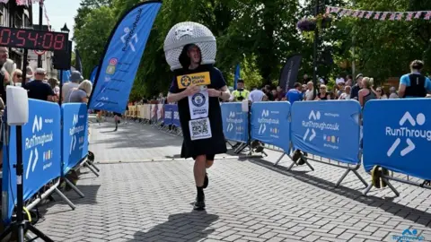 Dan Hudson Participant dressed in a large helmet-shaped costume runs along a race course toward the finish area, wearing a Marie Curie charity bib and official attempt sign. Blue event barriers, spectators, flags and a race clock line the route beneath trees on a sunny day.