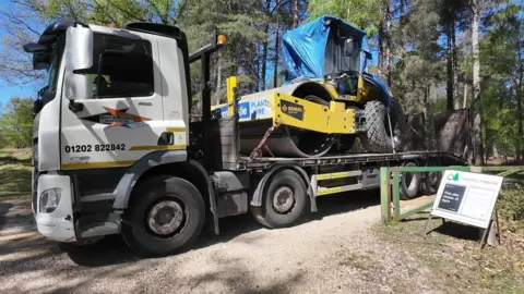 The road roller is on the back of a truck, covered in tarpaulin, which is driving away.