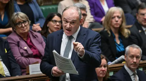 House of Commons Liberal Democrat leader Sir Ed Davey speaking during Prime Minister's Questions, with MPs sitting on green benches behind him. He is pointing with one hand and holding papers in the other.