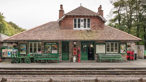 Woody Bay Station viewed from across the railway tracks. It is in the style of a Swiss chalet and has two chimneys, a slanted roof and green doors. There are green benches outside it and old-style luggage carrying trolleys. There is also a post box built in to the front wall. There are trees beyond.