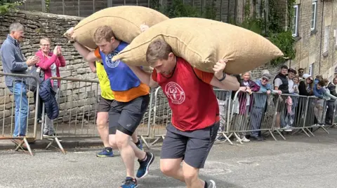 Three men wearing coloured bibs are running up a steep hill with a sack of wool on their backs. A crowd of people are watching them from behind a barrier.