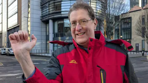 Jonathan Burns, wearing a red waterproof coat with grey sleeves, holding his hand up to the camera with a very small scar - no more than a centimetre - visible on his wrist. He has brown hair and round glasses. He is standing in Bedford Street, behind him is a modern high-rise building of glass and steel.