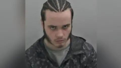 Zach Pearson in a police mugshot. He has braids in his hair and he has his head bowed but is looking at the camera.