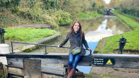 A woman sits on a wooden canal lock with the waterway visible behind her. She has long dark hair, and is wearing a grey jacket, blue jeams and brown leather boots.