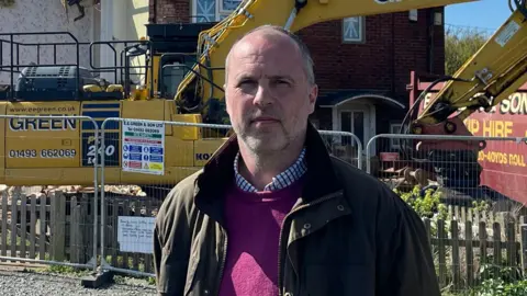 Edd Smith/BBC Rob Goodliffe standing in front of a digger at a house. There is also a red skip and metal fencing.