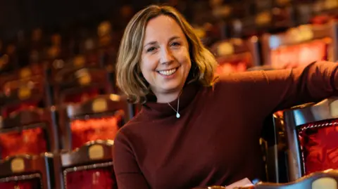 Theatre Royal Newcastle Newcastle Theatre Royal chief executive Marianne Locatori sitting in the theatre's red stalls. She has shoulder length blonde highlighted hair and is wearing a dark red polo neck and a necklace.