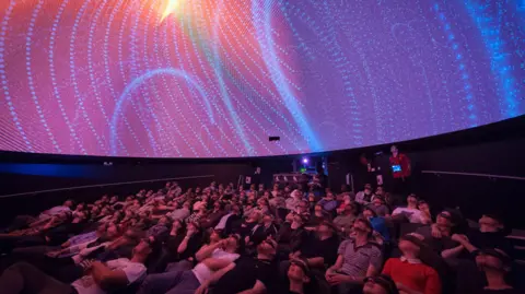 An audience looking up at the ceiling in a planetarium. The display is pink and there are blue dots all over it.