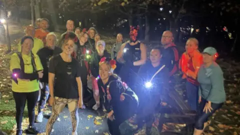 Abbie Blackburn The image shows a group of people gathered outdoors at night on a paved path surrounded by fallen leaves. Most individuals are wearing athletic or casual clothing, and several have headlamps or handheld lights, which are illuminating the scene. The group appears to be preparing for or finishing an activity, possibly a night run or walk. In the background, there are trees with autumn foliage.