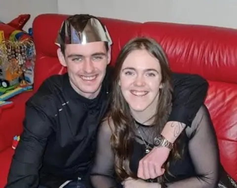 Margaret McCallum A young man and woman sitting on a red couch. The man is wearing a party hat and has his arm around the woman - both are smiling at the camera.