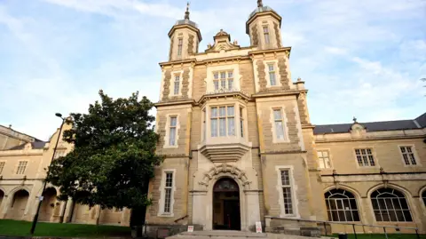 Sandy coloured exterior of Snaresbrook Crown Court building with tree to left of entrance