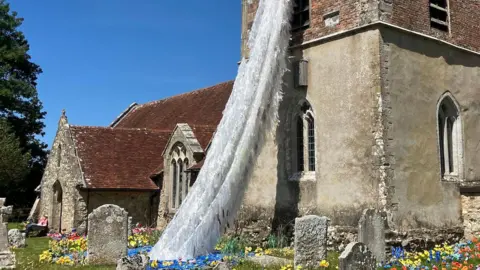 A picture of an ancient church. A cascade of white created to look like a waterfall is coming from the tower. It ends in a blue pool in the graveyard, there are colourful knitted flowers in amongst the gravestones. 