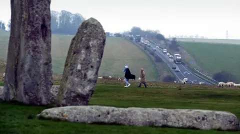 PA Media The A303 as seen from Stonehenge. Two walkers can be seen in the middle ground on a grey day.