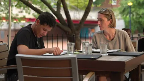 Rae and Lindsay sit at an outdoor restaurant table looking at menus. Rae has short dark hair and is wearing a black T-shirt. Lindsay has light brown hair in a bob and is wearing a beige T-shirt.
