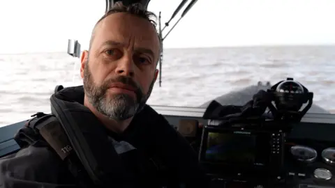 Inside the cabin of a police patrol boat, an officer in black protective gear sits near the control console. PC Mark Thompson has a brown beard and moustache, which is greying, and wears a black life jacket around his neck. Large windows wrap around the cabin, giving a wide view of the open water under an overcast sky.