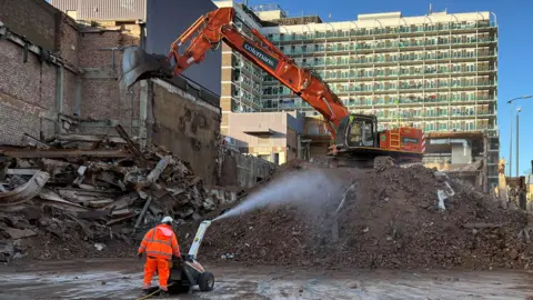 Ollie Conopo/BBC A pile of rubble with an orange digger on top, with a block of flats in the background.