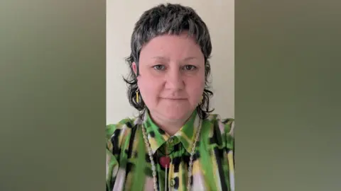 Supplied Susan has short dark hair and is wearing a green, yellow, black and white patterned shirt and a clear beaded necklace.