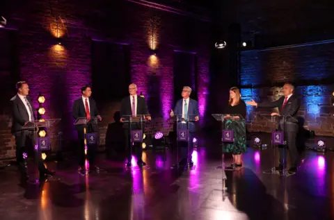 PA Media Six people stand at lecterns during a televised debate in a studio with purple and blue lighting against exposed brick walls; one speaker gestures toward another while the others look on.