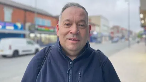 A man in a navy fleece stands on a high street. A van and shops can be seen blurred behind him. the man has short white hair and a half-smile on his face.