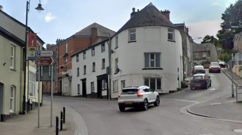 The top of St Nicholas Street street splits into two roads. There is a rounded three-storey building on the apex. To the left are signs indicating a long stay car park and the town museum and Shire Hall. 