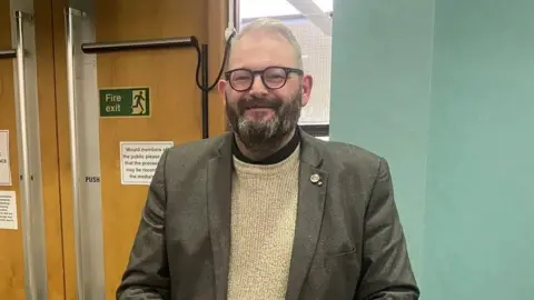 BBC Mike Young has a dark beard that is greying and grey hair. He wears glasses and smiles at the camera. He is wearing a brown suit jacket and beige jumper.