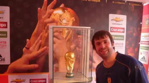 Graeme McNay Graeme McNay wearing a Scotland jersey, and standing next to the World Cup trophy, which is in a glass case.