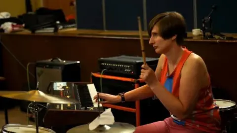 Mary McKay A woman with short brown hair wearing a red vest and trousers playing the drums.