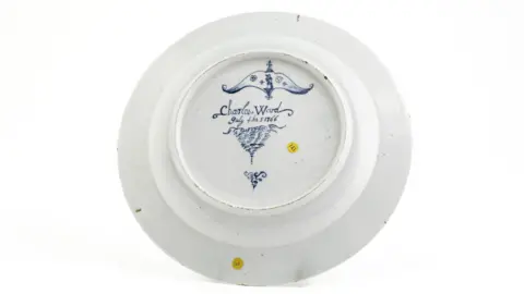 Woolley and Wallis A professional-looking close-up image of a white plate with the words "Charles Ward… July the 5 1766" inscribed on it in blue paint. It is propped up and pictured in front of an all white background.