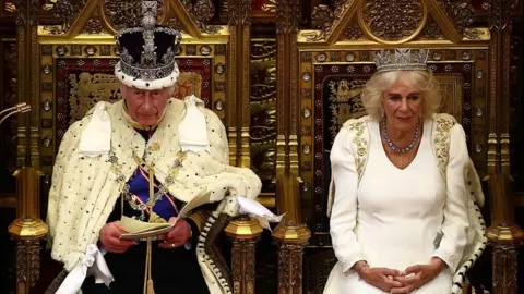 Getty Images Wearing the Imperial State Crown and ceremonial robes, King Charles delivers the 2024 King's Speech from the throne in the House of Lords. Queen Camilla, also wearing ceremonial robes, sits to his left, on the right of the picture.