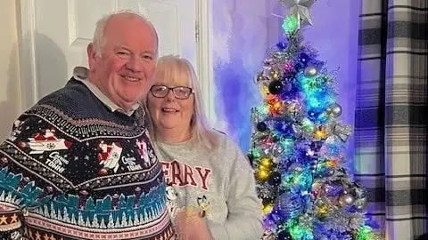 A middle-aged man and woman stand with their arms around each other. They are wearing Christmas jumpers and standing in front of a Christmas tree. 