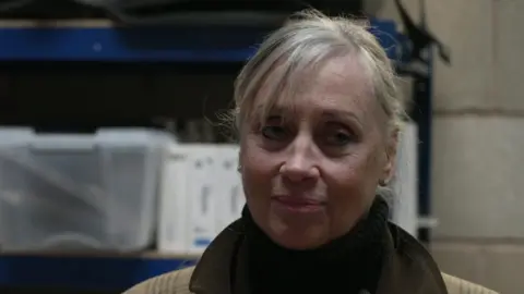 An older woman with short hair, pinned back, wearing a brown coat and pearl stud earrings, and pink lipstick, looks to the right of the camera 