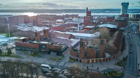Liverpool City Region Combined Authority An ariel view of how the new station might look from St James Place. The station is made of red brick and you can see the word Baltic in large letters along one wall. The Cains Brewery can be seen in the distance, and beyond that the river Mersey. 