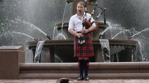 A man with a red kilt and white shirt playing the bagpipes in front of a fountain