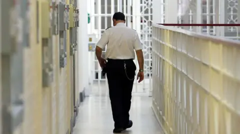 A prison officer walks down the corridor of a prison with his back to the camera. 